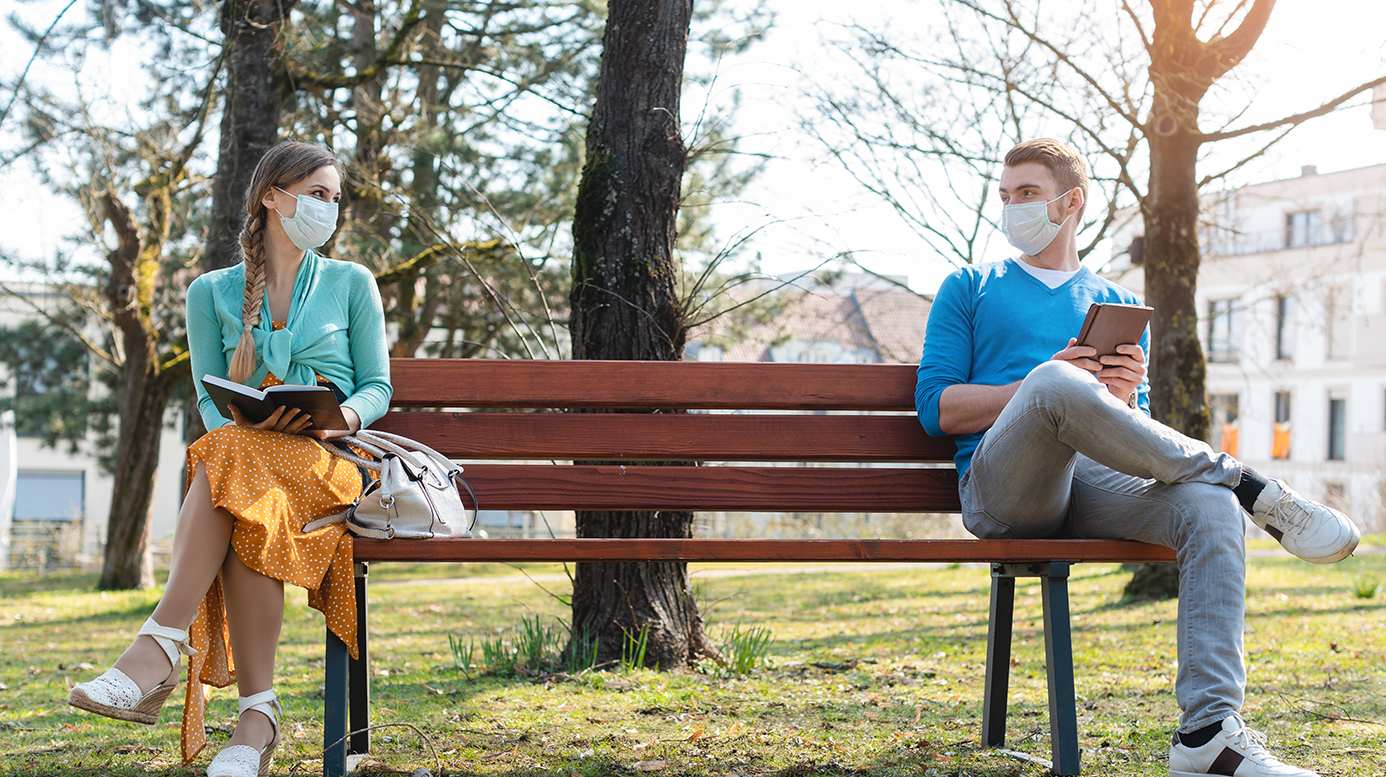 A young woman and a young man sitting wearing masks, sitting on opposite ends of a park bench
