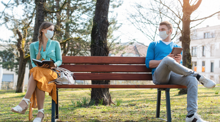 A young woman and a young man sitting wearing masks, sitting on opposite ends of a park bench